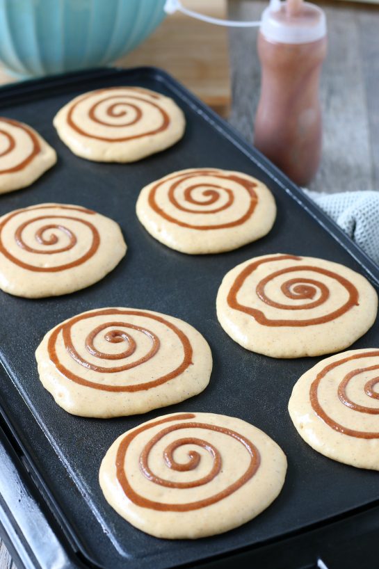 Eight cinnamon roll pancakes cooking on the hot griddle almost ready to eat.