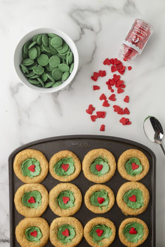 Adding the green melting chocolates and red hearts for the Grinch Cookie Cup Bites recipe 